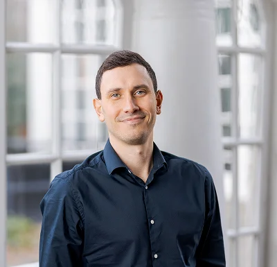 Business portrait of Alex Elsbury, smiling man in dark blue shirt, standing by large window in a bright office.