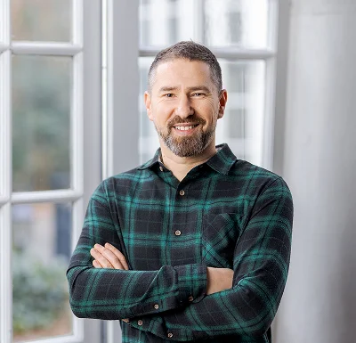 Business portrait of Alexandre Hachet, smiling bearded man in green check shirt, arms crossed by a window.