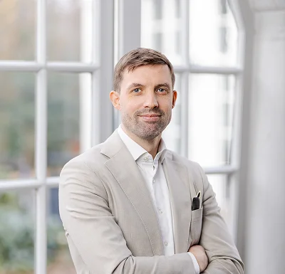 Business portrait of Jamie Duijn, smiling man in light suit with arms crossed, standing by a window.